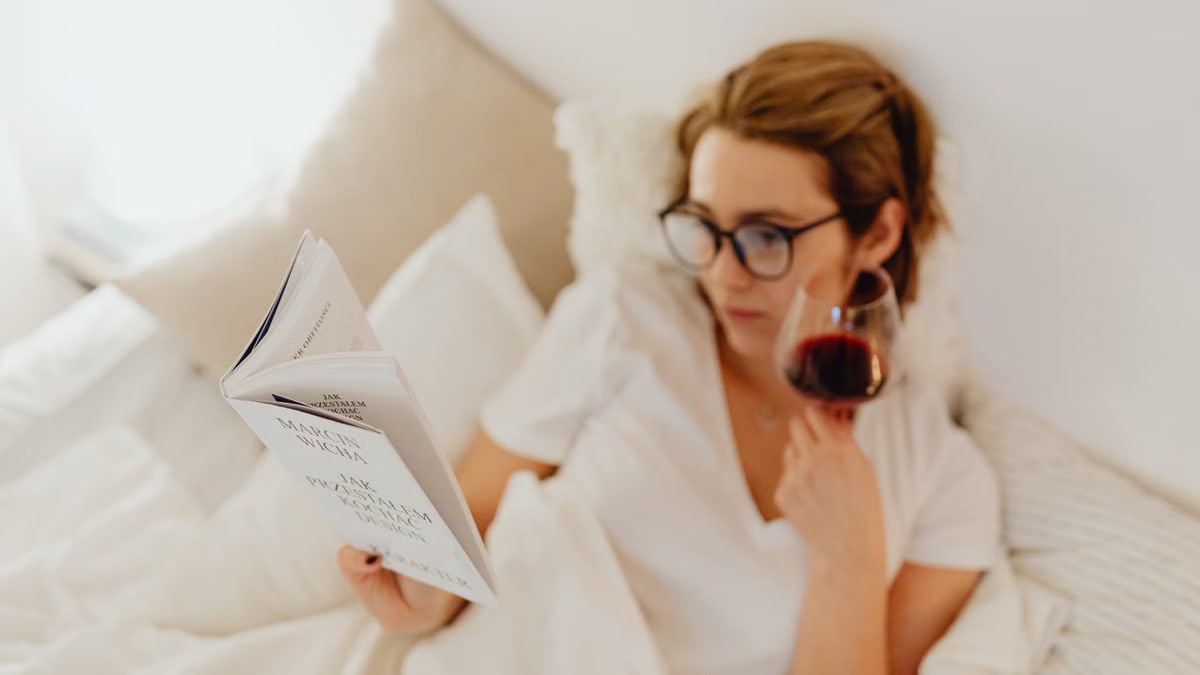 Woman enjoying relaxation in bed, reading a book with a glass of wine.